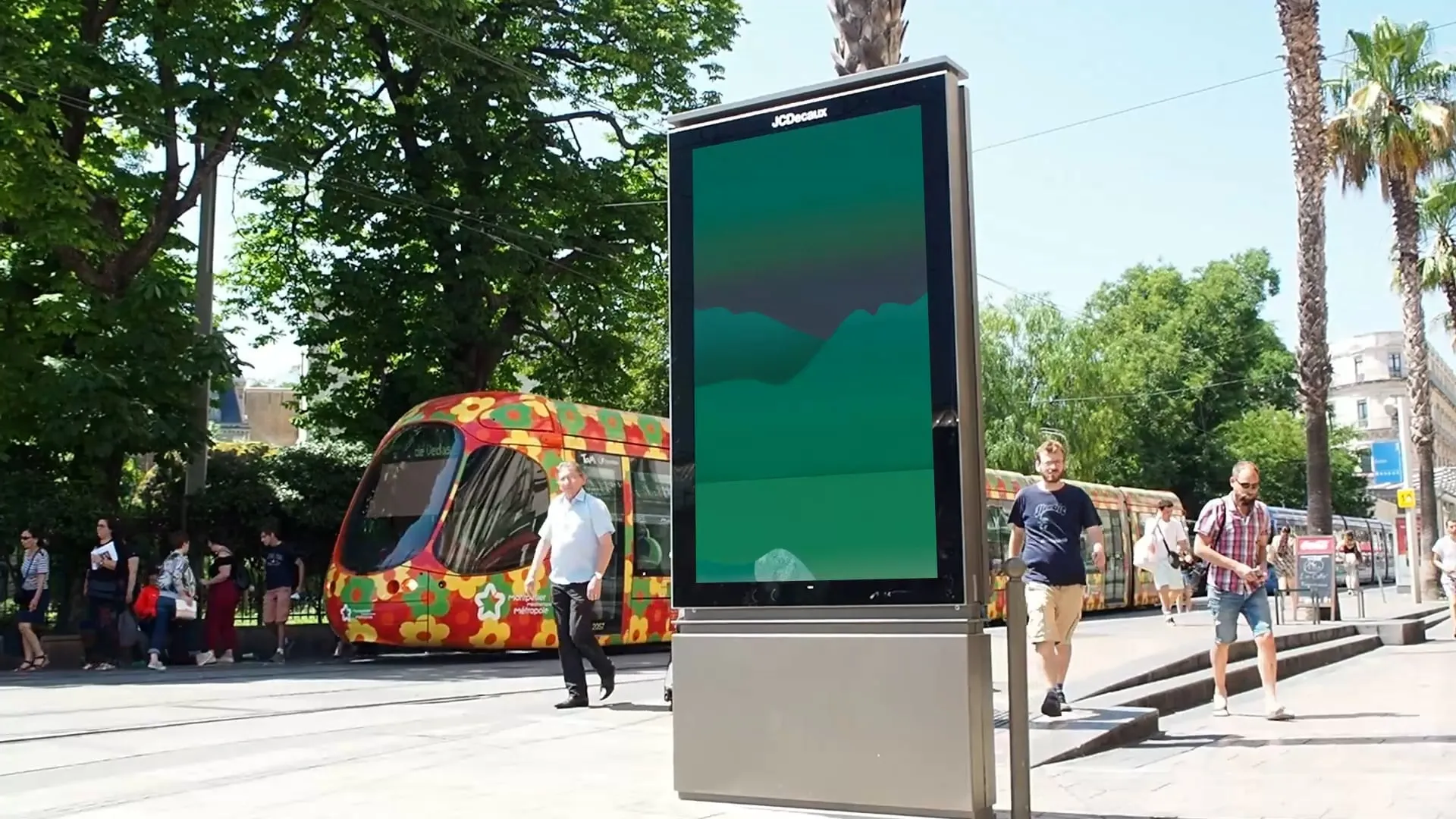 Un écran publicitaire sur un trottoir ensoleillé, un tramway coloré passe derrière, l'affiche animée à l'écran
